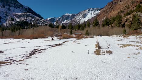 Aerial-Of-A-Woman-And-Her-Pet-Dog-Playing-Beside-A-Winter-Frozen-Lake-In-The-Sierra-Nevada-Mountains,-Near-Yosemite,-California