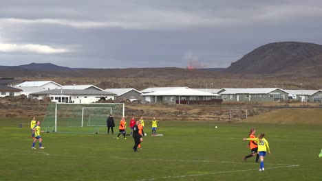 The-Fagradalsfjall-Volcano-Is-Seen-Erupting-In-The-Distance-Behind-A-Girls-Soccer-Match-The-Town-Of-Grindavik,-Iceland