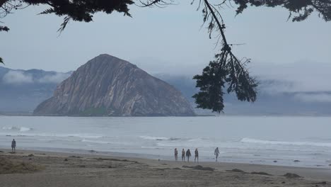 People-Walk-On-A-Beach-Near-Morro-Bay,-California-With-Morro-Rock-In-Distance