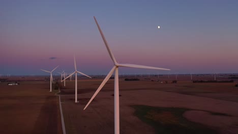 Excelente-Antena-Al-Atardecer-De-Molinos-De-Viento-Que-Generan-Electricidad-Limpia-En-Las-Tierras-De-Cultivo-Planas-De-Indiana,-Illinois,-Con-La-Luna-Saliendo