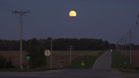 Un-Automóvil-Distante-Conduce-Por-Una-Carretera-Rural-Solitaria-Por-La-Noche-Con-La-Luna-Llena-Saliendo