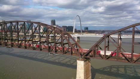 Antena-De-Un-Tren-De-Carga-Que-Cruza-El-Río-Mississippi-Con-El-Arco-De-La-Puerta-De-Enlace-De-St-Louis-Y-El-Fondo-Del-Horizonte-De-La-Ciudad