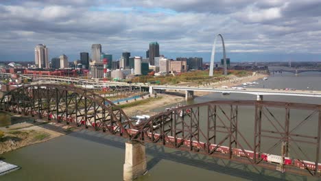 Antena-De-Un-Tren-De-Carga-Que-Cruza-El-Río-Mississippi-Con-El-Arco-De-La-Puerta-De-Enlace-De-St-Louis-Y-El-Fondo-Del-Horizonte-De-La-Ciudad