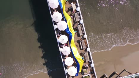 Manifestantes-Ucranianos-Aéreos-De-Arriba-Hacia-Abajo-Con-Una-Gran-Bandera-Ucraniana-Protestan-Contra-La-Invasión-Rusa-De-Su-País,-En-El-Muelle-De-Malibu,-California