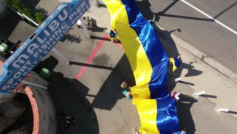 Manifestantes-Ucranianos-Aéreos-De-Arriba-Hacia-Abajo-Con-Una-Gran-Bandera-Ucraniana-Protestan-Contra-La-Invasión-Rusa-De-Su-País,-En-El-Muelle-De-Malibu,-California-Con-El-Cartel-De-Malibu