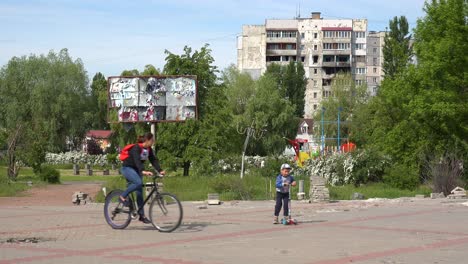 Una-Mujer-Monta-Una-Bicicleta-Con-Su-Hijo-Con-La-Ciudad-Destruida-De-Bodyanka,-Ucrania,-Antecedentes