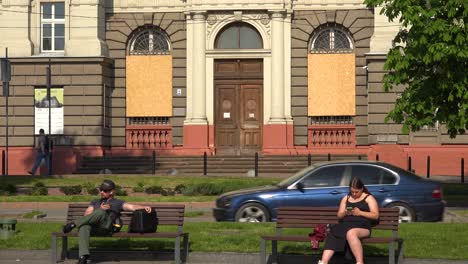 Los-Ucranianos-Se-Sientan-En-Un-Banco-Del-Parque-Disfrutando-Del-Sol-En-El-Centro-De-Lviv,-Ucrania-Con-Edificios-Tapiados-En-El-Fondo