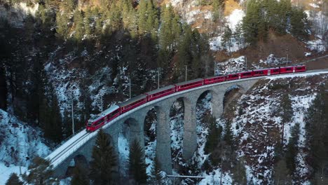 Excelente-Vista-Aérea-De-Un-Tren-Que-Viaja-A-Través-Del-Puente-Una-Región-Montañosa-Nevada-De-Suiza