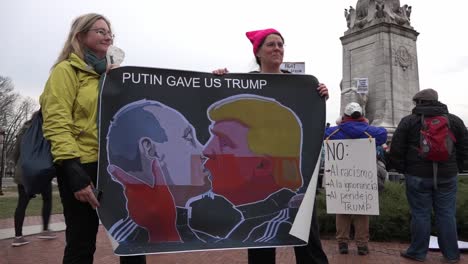 Two-Female-Political-Protestors-Carry-A-Sign-Of-Donald-Trump-And-Vladmir-Locked-In-An-Embrace,-Washington-Dc