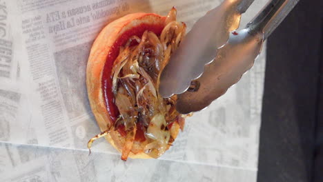 Chef-placing-grilled-onion-with-tongs-on-hamburger-bun-with-sauce