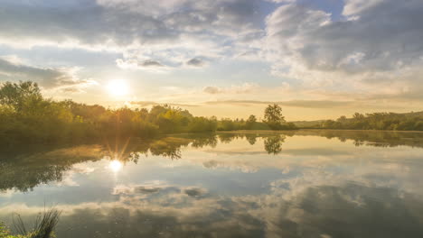 Time-lapse-capture-on-the-pond-at-the-morning-sunrise-over-trees-lying-near-water-and-moving-clouds-over-water-with-reflection-on-water-surface