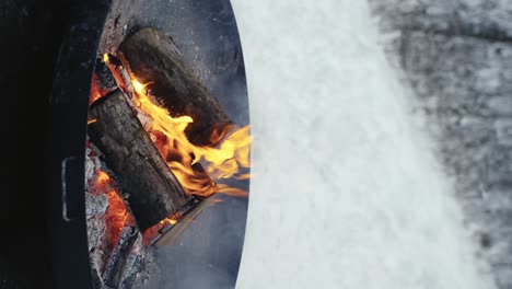 Epic-4K-vertical-shot-of-wood-logs-burning-inside-a-brazier-while-it-snows-outside