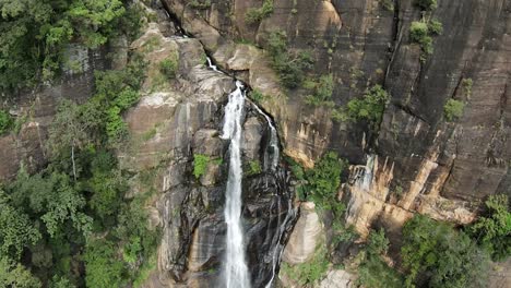 Aerial-dolly-in-over-Ravana-falls-streaming-down-rocky-vertical-cliff-covered-in-green-vegetation,-Sri-Lanka