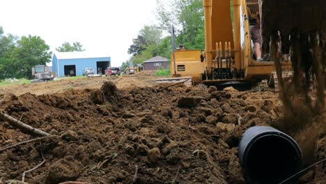 Tiefwinkelansicht-Eines-Mannes,-Der-Einen-Gelben-John-Deere-Bagger-Bedient,-Um-Ein-Entwässerungsrohr-Für-Einen-Teich-An-Einem-Neuen-Landentwicklungsstandort-Abzudecken