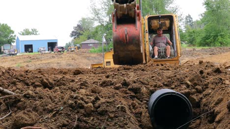 Tiefwinkelansicht-Eines-Mannes,-Der-Einen-Gelben-John-Deere-Bagger-Bedient,-Um-Ein-Entwässerungsrohr-Für-Einen-Teich-An-Einem-Neuen-Landentwicklungsstandort-Abzudecken