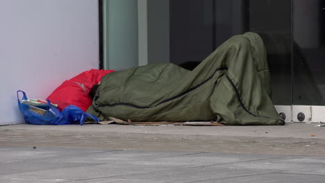 In-slow-motion-people-walk-past-a-homeless-person-sleeping-in-the-doorway-of-an-empty-retail-space,-known-as-“dead-spaces”,-on-Tottenham-Court-Road