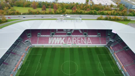 Vuelo-Aéreo-Sobre-La-Azotea-Del-Estadio-Vacío-Wwk-Arena,-Hogar-Del-Fc-Augsburg-Con-La-Carretera-De-Doble-Calzada-B17-En-El-Fondo