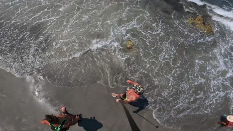 High-angle-top-down-view-of-man-with-cap-walking-on-crowded-beach-shore-enjoying-hot-summer-day-while-holding-long-camera-pole