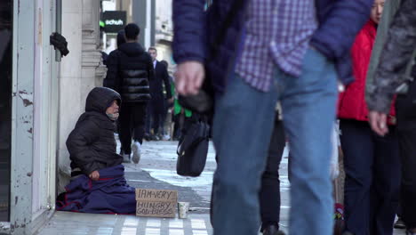 Leute-Gehen-An-Einem-Obdachlosen-Vorbei,-Der-Auf-Der-Oxford-Street-Sitzt-Und-Um-Geld-Bettelt,-Mit-Einem-Pappschild,-Auf-Dem-Steht:-„Ich-Bin-Sehr-Hungrig,-Gott-Segne-Dich“