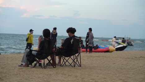 Pareja-Sentada-Junto-A-La-Playa-Y-Disfrutando-De-La-Vista-Al-Mar-Mientras-Revisa-Sus-Teléfonos-En-Pattaya,-Tailandia