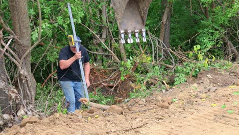 A-man-with-surveyor's-tools-checks-the-depth-of-an-overflow-drainage-ditch-of-a-pond-on-the-edge-of-a-land-development-and-construction-site