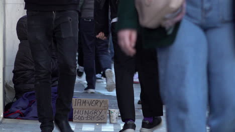 Leute-Gehen-An-Einem-Obdachlosen-Vorbei,-Der-Auf-Der-Oxford-Street-Sitzt-Und-Um-Geld-Bettelt,-Mit-Einem-Pappschild,-Auf-Dem-Steht:-„Ich-Bin-Sehr-Hungrig,-Gott-Segne-Dich“