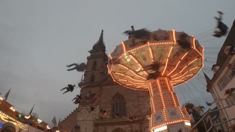 Gente-Montando-El-Carrusel-De-Cadena-Frente-A-La-Catedral-En-La-Feria-De-Otoño-En-Basilea,-Suiza