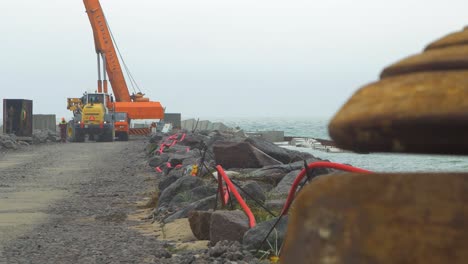 Sitio-De-Construcción-En-La-Costa-Del-Mar-Báltico,-Fortalecimiento-De-La-Costa-Del-Mar-Báltico,-Construcción-De-Un-Muelle-Protector-De-Piedra,-Maquinaria-Pesada-Descargando-Bloques-De-Hormigón,-Día-Nublado