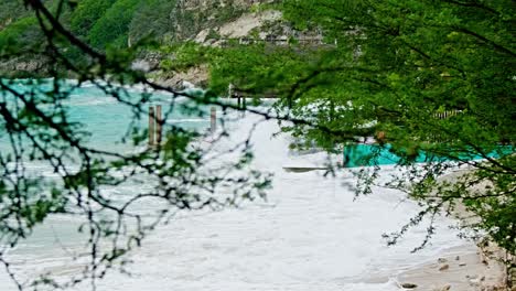 Zoom-shot-with-foliage-foreground-of-local-wooden-fishing-boat-being-smashed-into-land-during-sudden-storm-with-rough-waves,-Caribbean