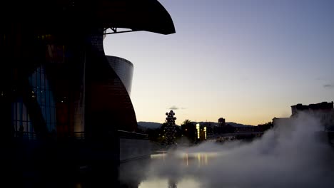 Museo-Guggenheim-Con-Niebla-De-Agua-Y-árbol-Alto-Y-La-Escultura-Del-Ojo-De-Anish-Kapoor-Afuera-Durante-La-Puesta-Del-Sol,-Plano-General