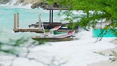 Slowmo-zoom-shot-with-foliage-foreground-of-local-wooden-fishing-boat-being-smashed-into-land-during-sudden-storm-with-rough-waves,-Caribbean