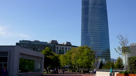 Electric-utility-Iberdrola-Tower-base-with-people-walking-promenade-below-it,-Wide-handheld-shot