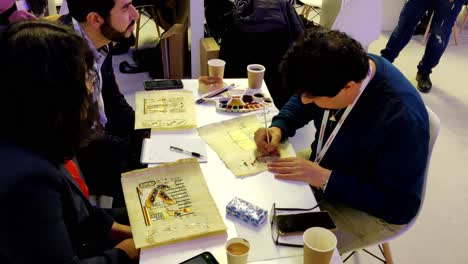 three-people-sitting-on-a-table-drawing-and-coloring-ancient-art-on-paper