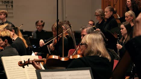 Medium-handheld-slow-motion-shot-of-violin-player-with-wooden-violins,-flute-player-and-choir-singers-in-orchestra-during-concert-to-mozart