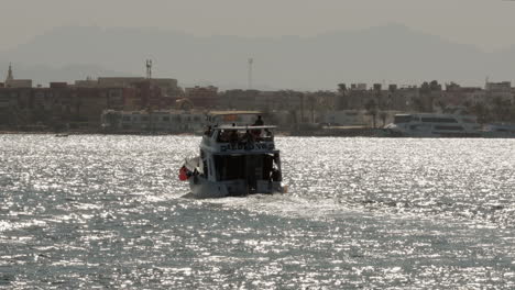 Un-Yate-Navegando-En-El-Mar-Hacia-La-Costa-En-El-Día-Soleado-De-Egipto,-Resplandor-Solar-En-La-Superficie-Del-Agua-Y-Paisaje-Urbano-Costero-En-El-Horizonte,-Paisaje-Costero