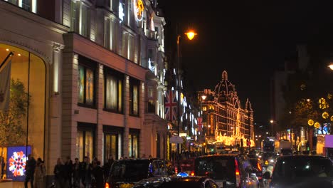 Toma-Panorámica-De-La-Famosa-Carretera-Brompton-Con-Los-Grandes-Almacenes-Harrods-Decorados-Para-Navidad-Con-Una-Acera-Abarrotada-Y-Un-Intenso-Movimiento-De-Tráfico-Por-La-Noche