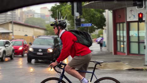 Hombre-Australiano-Cruzando-La-Carretera-En-Bicicleta-Con-Un-Casco-De-Diseño-único-Con-Bridas-Para-Disuadir-A-Un-Pájaro-Urraca-En-Un-Día-Lluvioso-En-Bowen-Hills,-Ciudad-De-Brisbane,-Queensland,-Australia