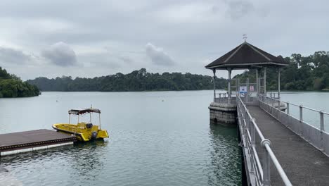 Tranquilidad-Del-Flujo-Tranquilo-Frente-A-La-Vista-De-Un-Pequeño-Bote-Amarillo-Y-Un-Pabellón-En-El-Embalse-Macritchie,-Singapur