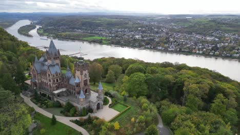 Toma-Panorámica-Del-Castillo-De-Drachenburg-En-Las-Colinas-De-Drachenfels-En-Königswinter-Con-El-Río-Rhein-Al-Fondo-En-Un-Fresco-Día-De-Otoño