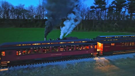 Un-Dron-Ligeramente-Por-Delante-Y-Una-Vista-Nocturna-Paralela-De-Un-Tren-De-Pasajeros-De-Vapor-Se-Detuvo-Cuando-Otro-Tren-De-Vapor-Pasa-Soplando-Mucho-Humo-Viendo-Las-Luces-En-Los-Vagones