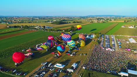 Una-Vista-Aérea-De-Varios-Globos-Aerostáticos-Que-Se-Lanzan-Al-Cielo-Durante-Un-Festival-Con-Multitudes-Observando,-En-Un-Día-De-Verano