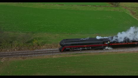 Vista-Paralela-De-Drones-De-Un-Tren-De-Pasajeros-De-Vapor-Que-Sopla-Humo-Y-Vapor-Cruzando-Un-Cruce-Con-Espectadores-Mirando-En-Un-Día-Soleado-De-Otoño