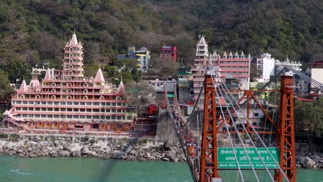 Antiguo-Templo-Hindú-En-La-Orilla-Del-Río-Ganges-Con-Un-Puente-De-Hierro-Que-Conduce-La-Línea-En-El-Día-Desde-Un-Video-De-ángulo-Plano-Tomado-En-El-Templo-Trimbakeshwar-Lakshman-Jhula-Rishikesh-Uttrakhand-India-El-15-De-Marzo-De-2022