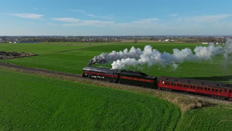 Un-Dron-Paralelo-A-La-Vista-Trasera-De-Un-Tren-De-Pasajeros-De-Vapor-Que-Sopla-Mucho-Humo-Y-Vapor-En-Un-Día-Soleado-De-Otoño