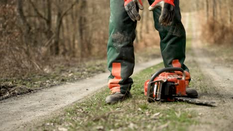 Tiro-De-ángulo-Bajo-A-Nivel-Del-Suelo-De-Arborist-Recogiendo-Motosierra-Y-Caminando-Por-El-Camino-Del-Bosque