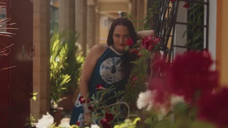 Beautiful-Indian-model-posing,-leaning-on-a-house-in-the-colorful-Fontainhas,-India,-wearing-a-blue-dress,-behind-bright-red-flowers