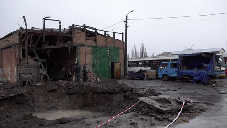 Arbeiter-Gehen-An-Einem-Russischen-S-300-raketenkrater-Vorbei,-Der-Sich-Mit-Regenwasser-Gefüllt-Hat,-Vor-Einer-Zerstörten-Garage-Und-Fahrzeugen-Auf-Einem-Busdepot