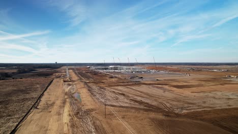 Acercamiento-Aéreo-Lento-De-Una-Gran-Estructura-De-Acero-En-Construcción-A-Lo-Lejos-Rodeada-De-Grúas,-Equipo-De-Construcción-Y-Tierra-Clasificada