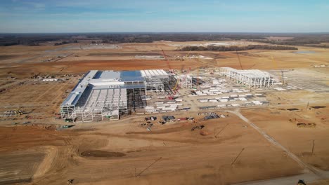 Alejamiento-Lento-Aéreo-Y-Desplazamiento-Panorámico-Alrededor-De-Una-Gran-Estructura-De-Acero-En-Construcción-Rodeada-De-Grúas,-Equipo-De-Construcción-Y-Tierra-Graduada