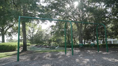 Leere-Schaukeln-Auf-Dem-Spielplatz-Mit-Sonne,-Die-An-Einem-Sommertag-Durch-Die-Bäume-Im-Hintergrund-Scheint